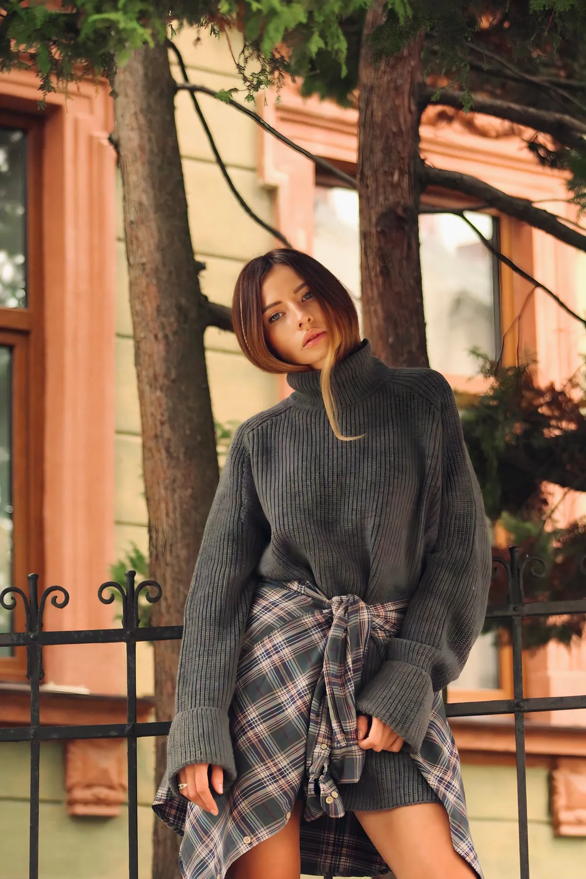 A woman wearing a sweater outdoors near a fence and greenery.