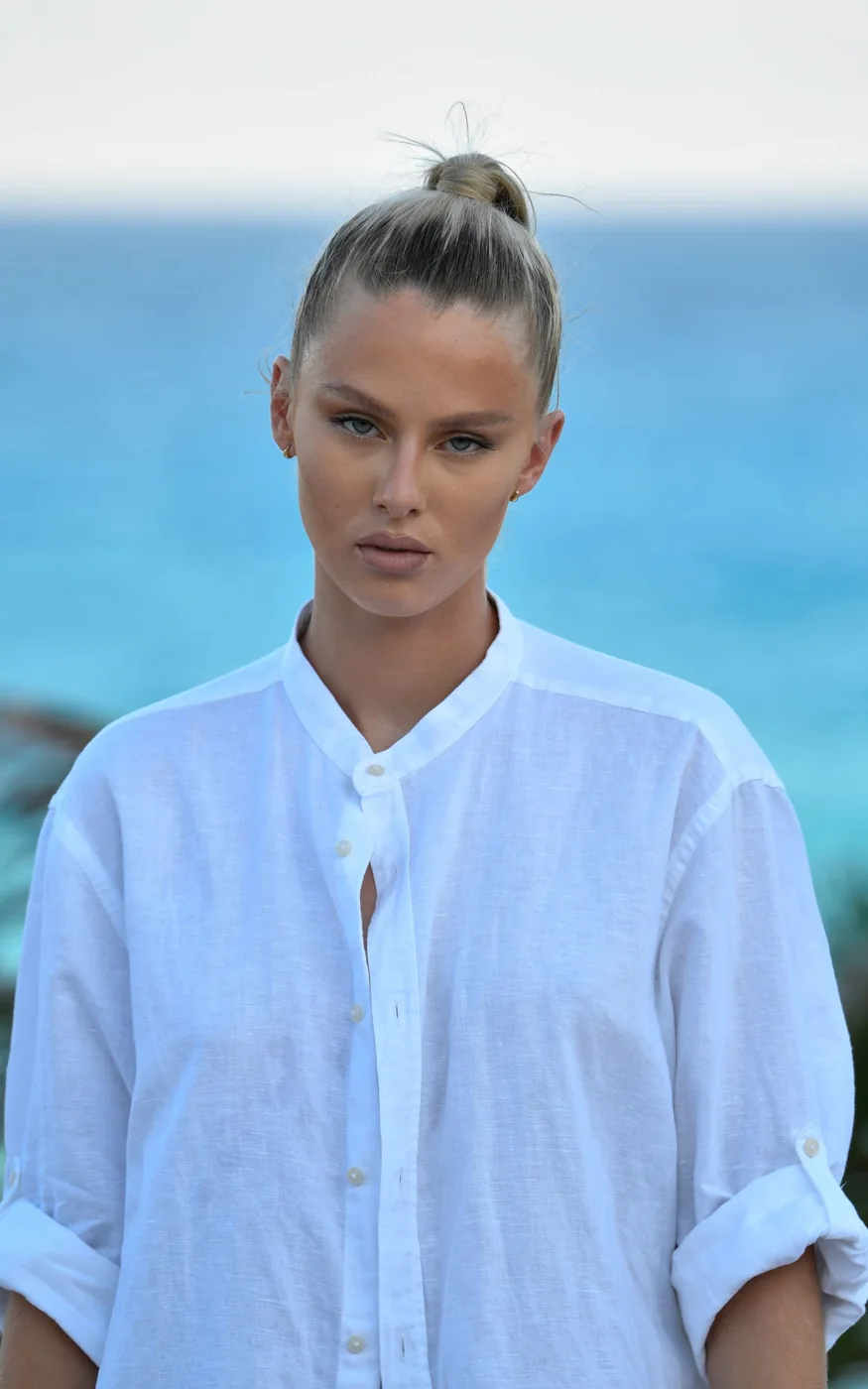 A woman in a pale band-collar shirt beside the sea.