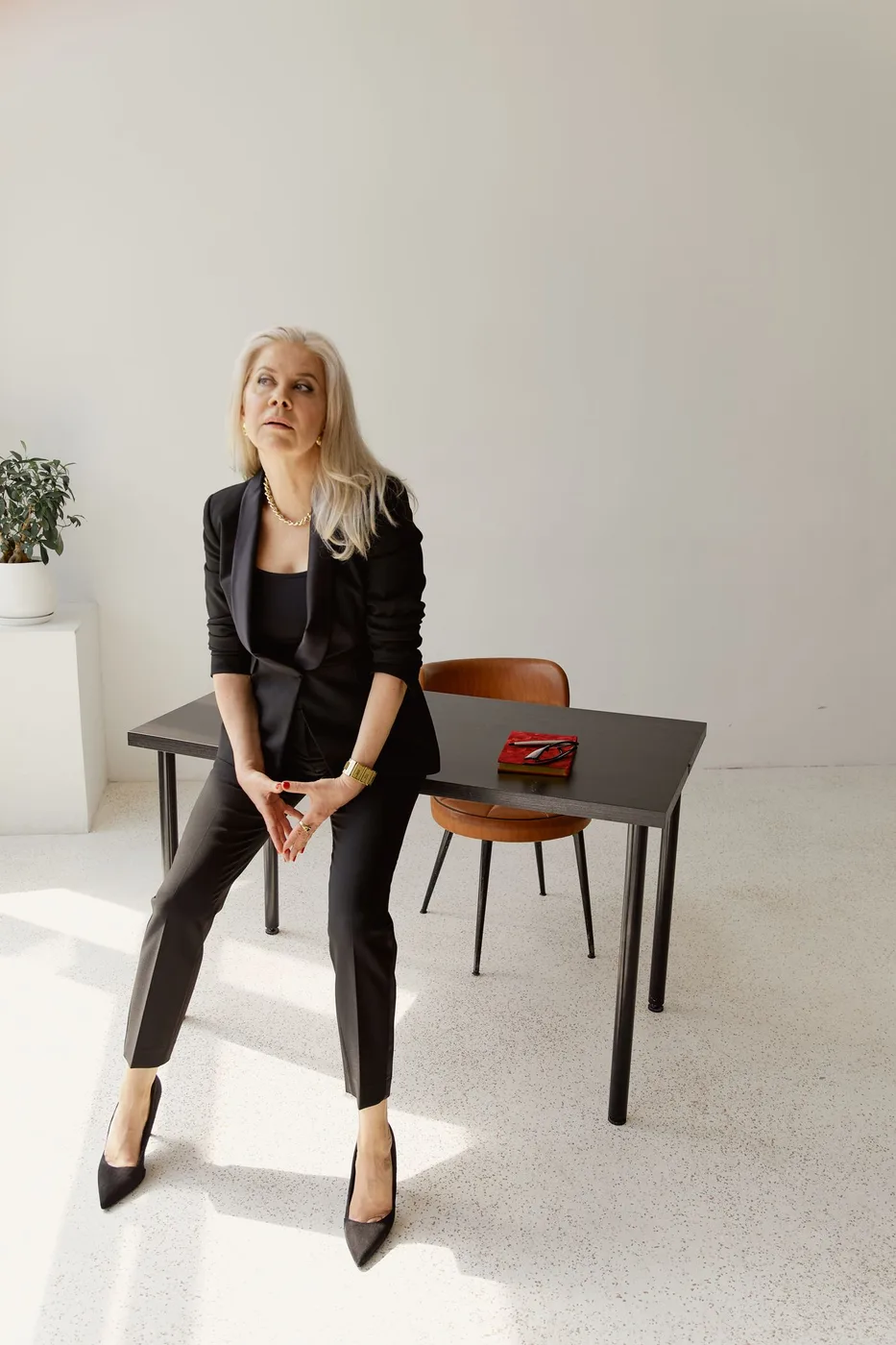 A woman wearing black tailoring seated indoors beside a small table.