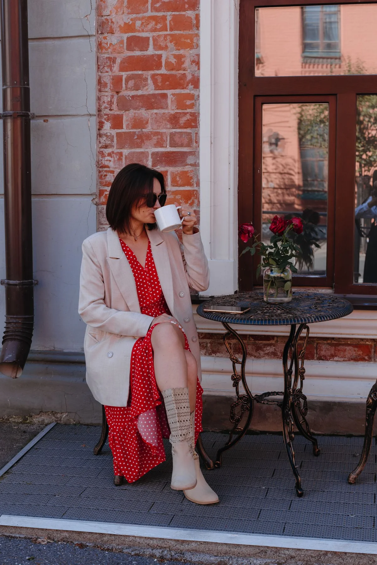 A person in a pale jacket and red outfit holding a cup beside a cafe.