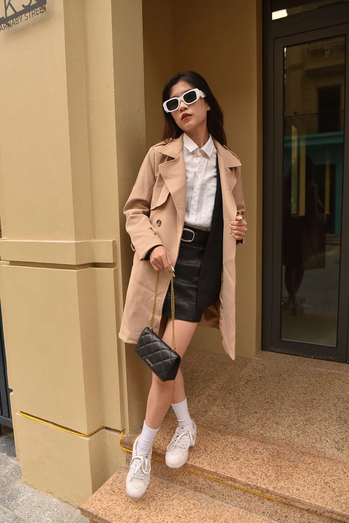 A woman in a pale coat and sunglasses at a building entrance.
