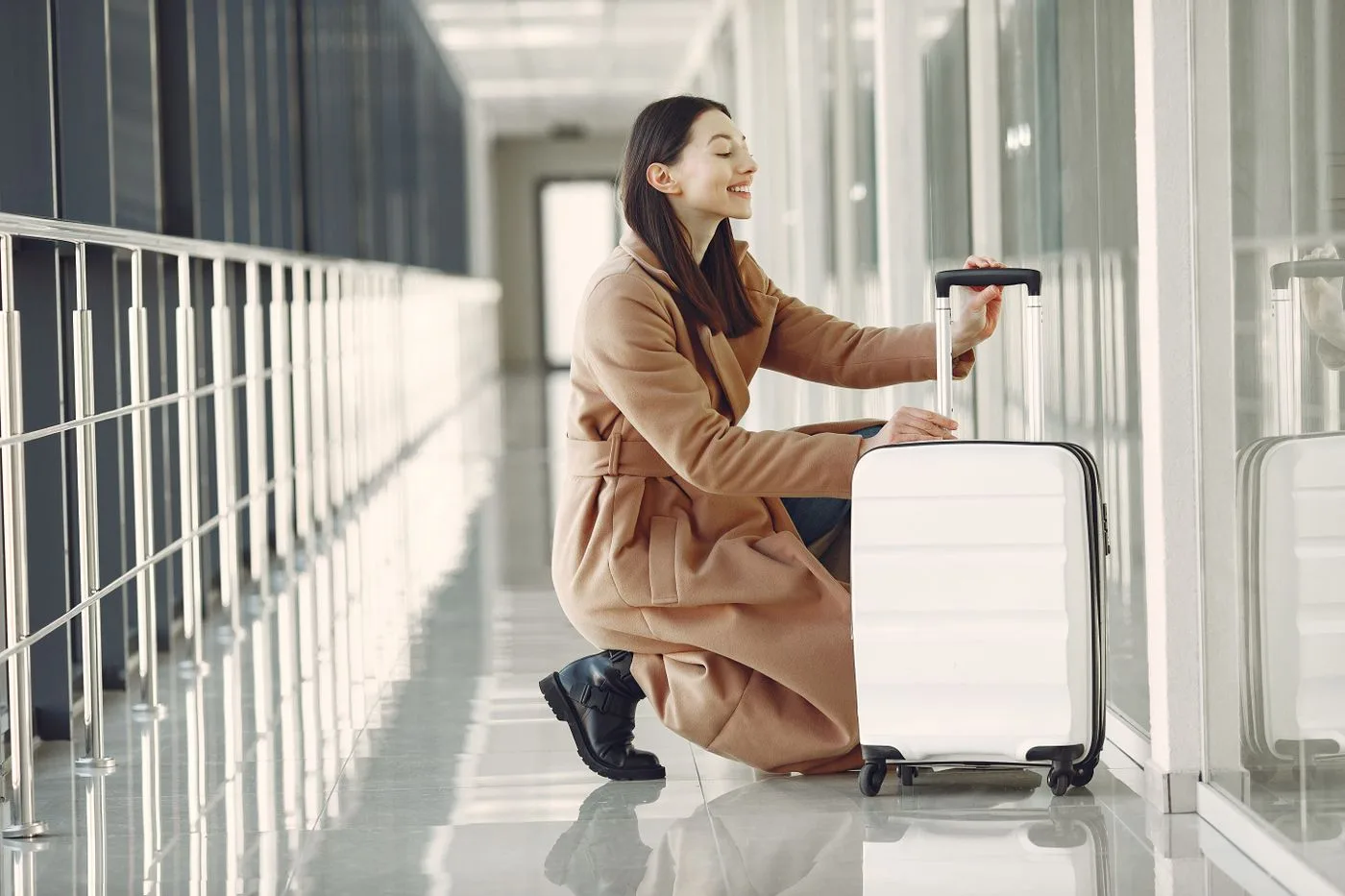 A woman kneeling beside a rolling suitcase in a corridor.