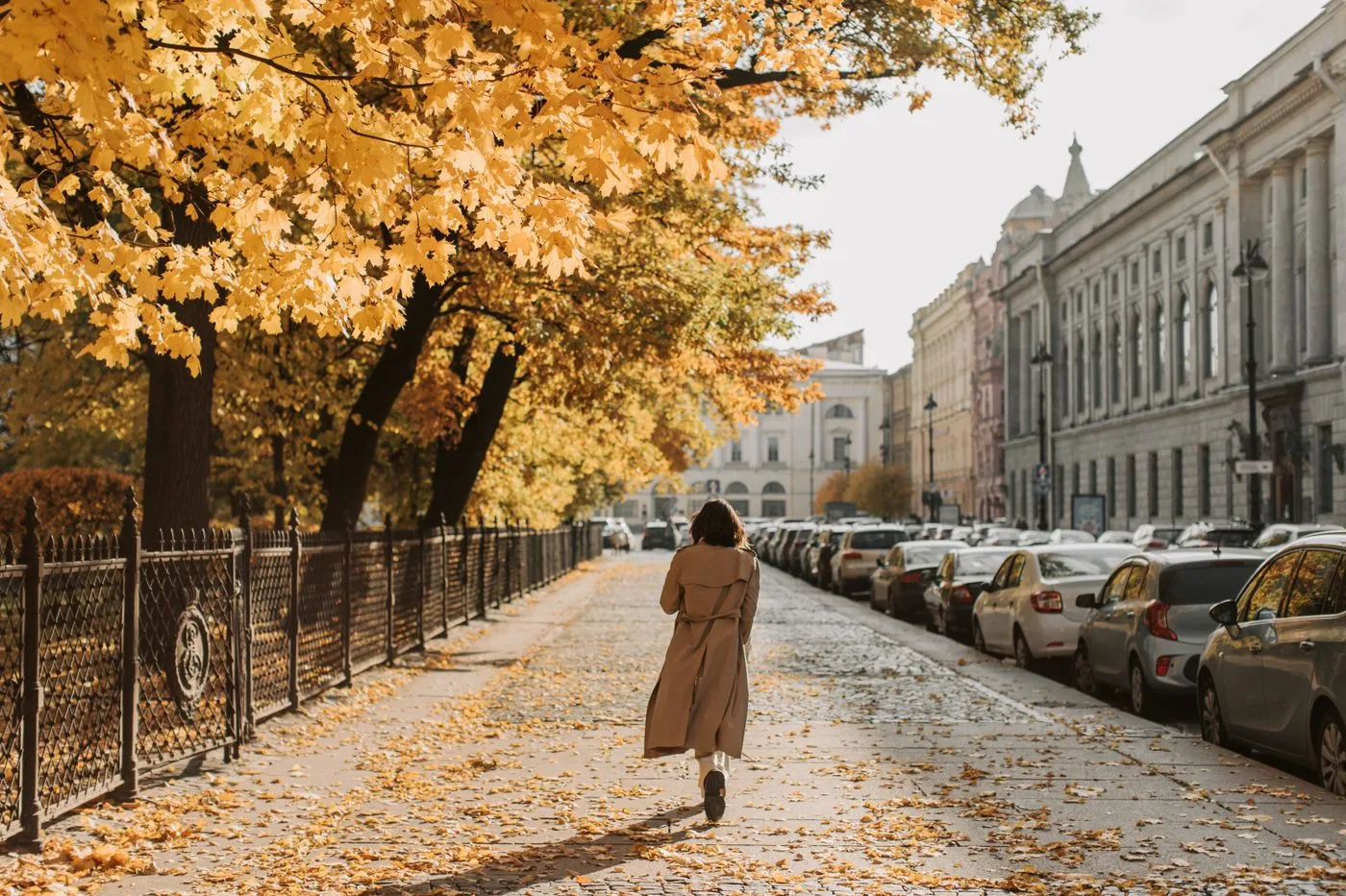 A person in a long brown coat on a leaf-covered city pavement.