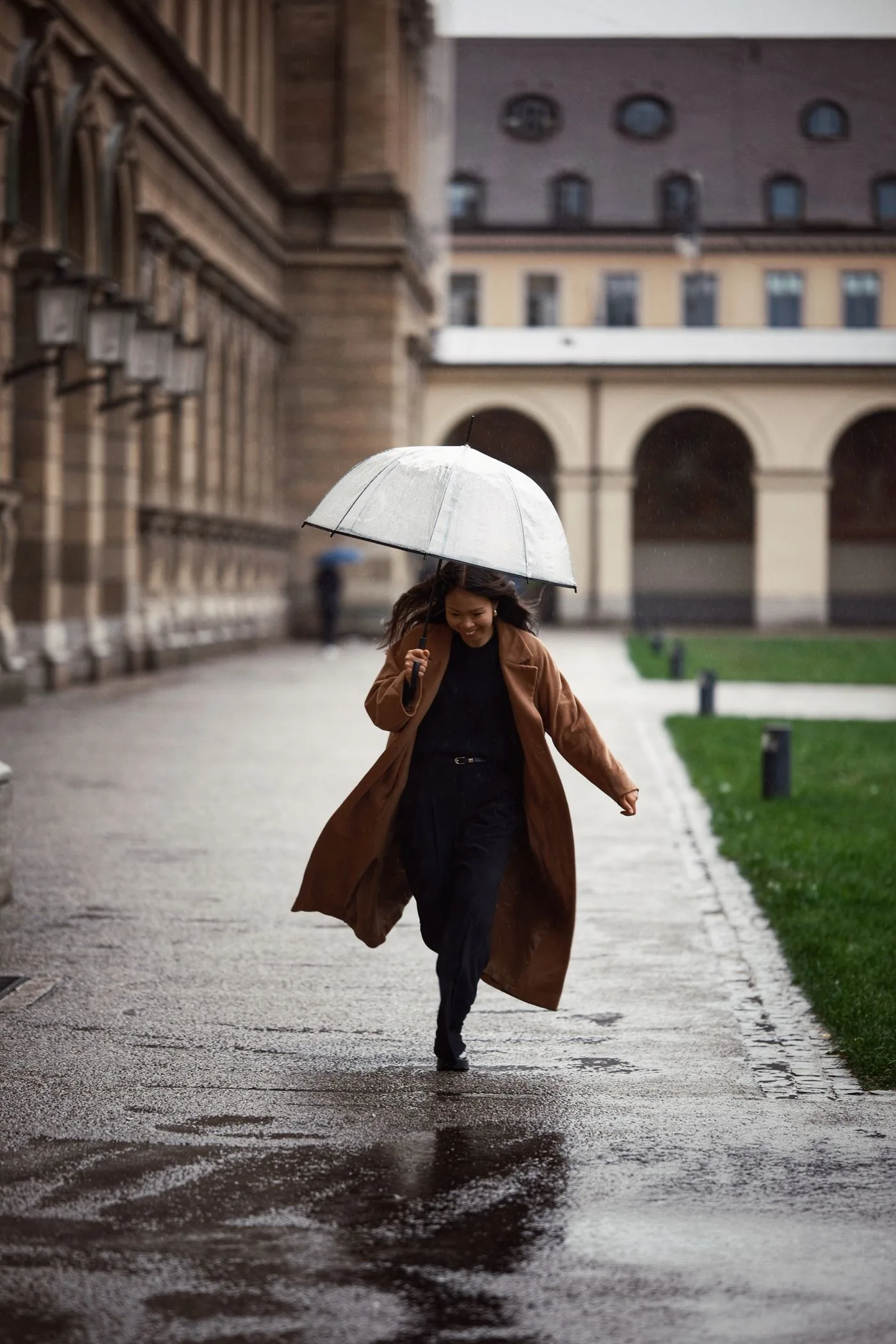 A woman in a brown coat carrying an umbrella on a wet pavement.