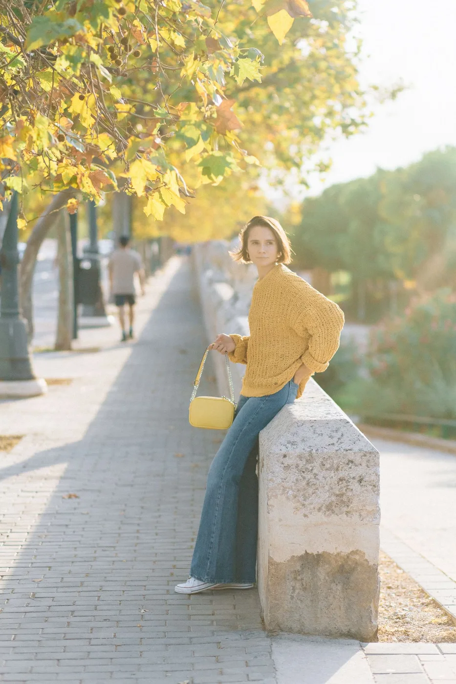 A woman wearing a yellow knit top and blue jeans on a sunlit street.